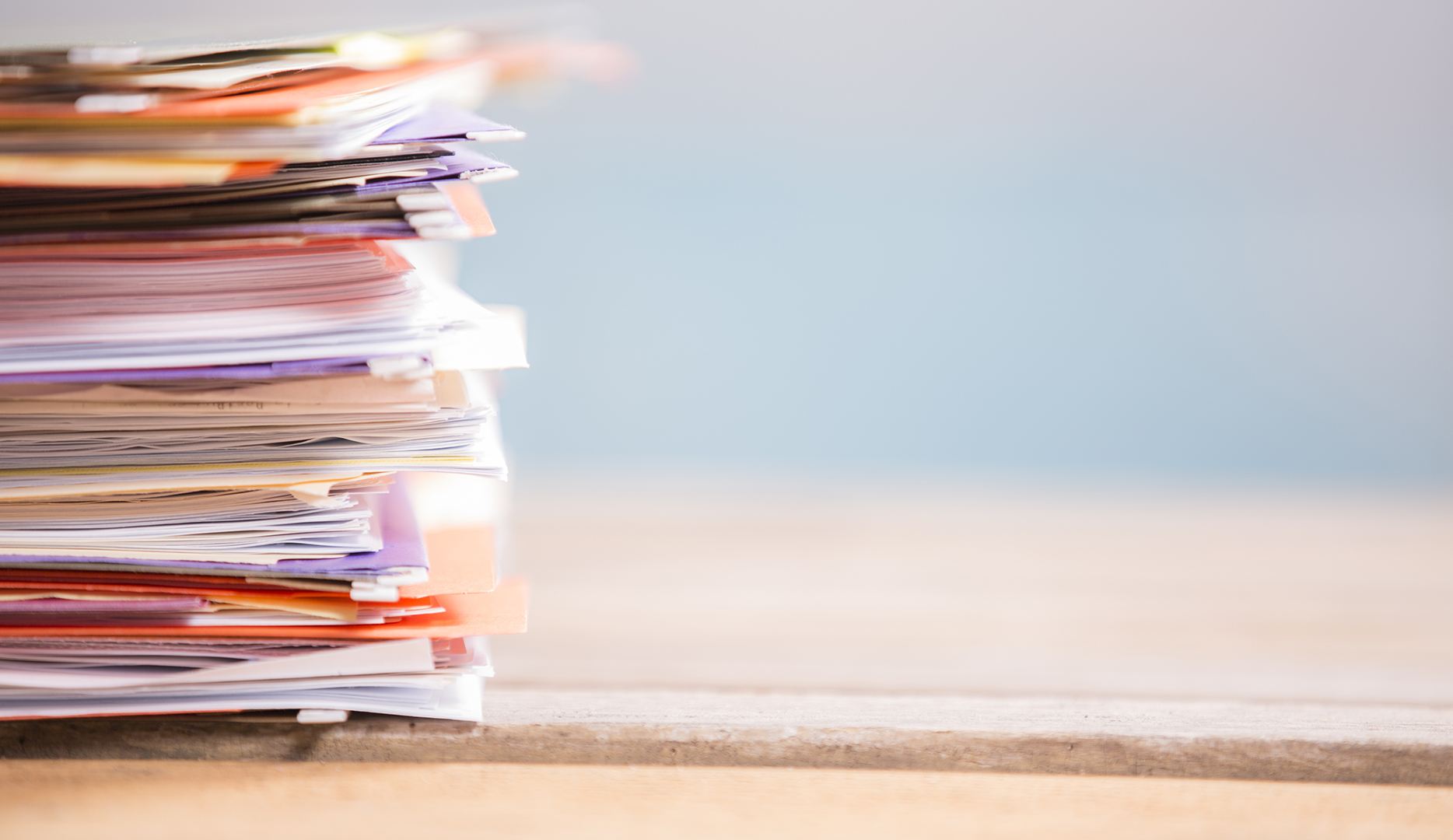 Large stack of multi-colored files and paperwork on office desk.  Blue window background. No people.